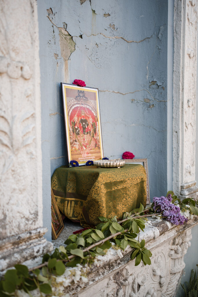 A traditional Hindu shrine set up with a picture of a deity, surrounded by flowers and ritualistic items, against a blue distressed wall.