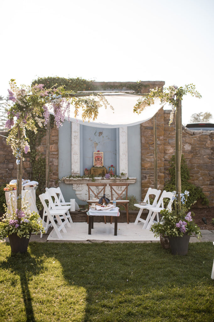 An outdoor Hindu wedding setup with a white canopy and floral decorations around a traditional shrine against a stone wall.