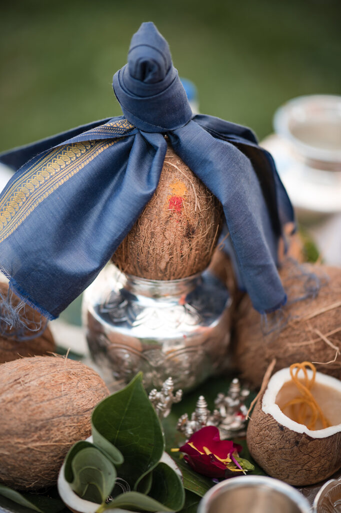 Close-up of a traditional Indian wedding ritual setup with a coconut wrapped in a blue cloth with golden details, placed atop a silver pot amidst an array of ceremonial items.