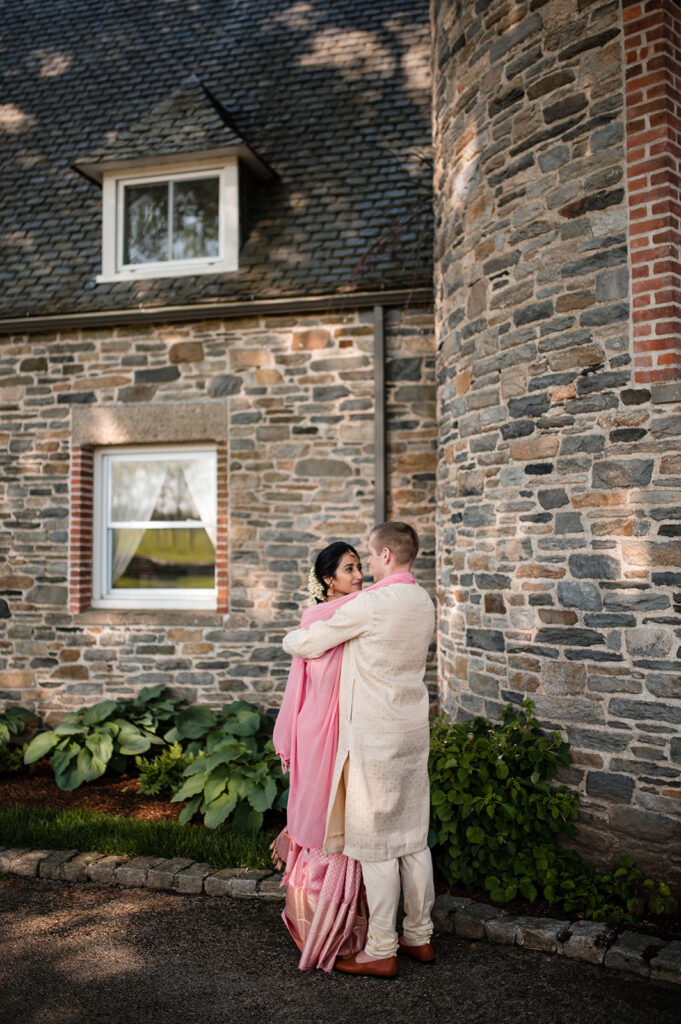 A bride and groom in traditional Indian wedding attire share a moment, with the bride in a pink saree, embraced by the groom in a cream sherwani, standing against an old brick building.