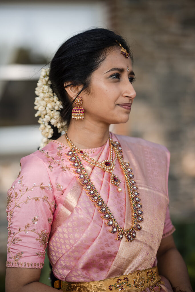 Portrait of a smiling Indian bride in a pink saree, adorned with elegant gold jewelry and a jasmine flower garland in her hair, gazing to the side.