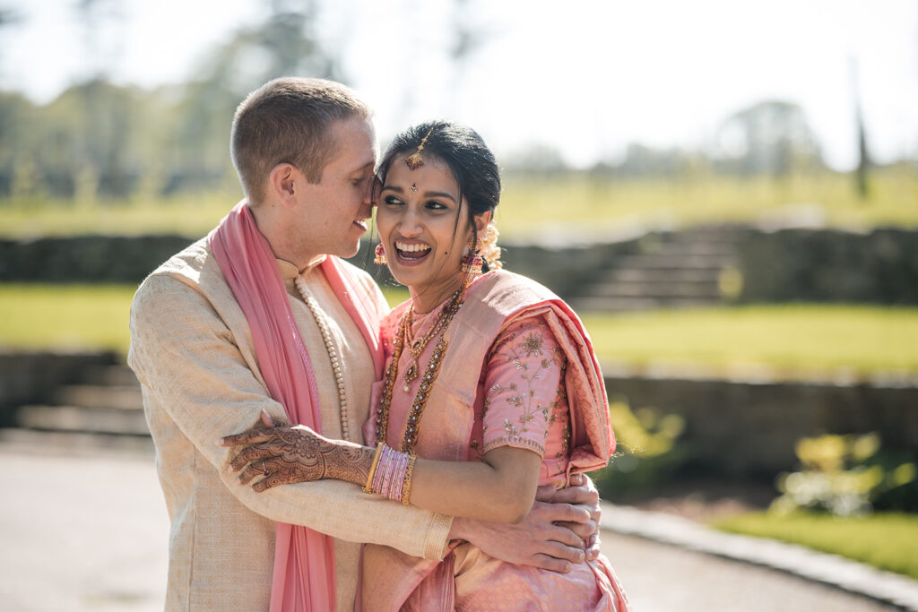 A joyous bride and groom outdoors, with the groom whispering into the bride's ear, making her laugh; both are in traditional Indian wedding attire.