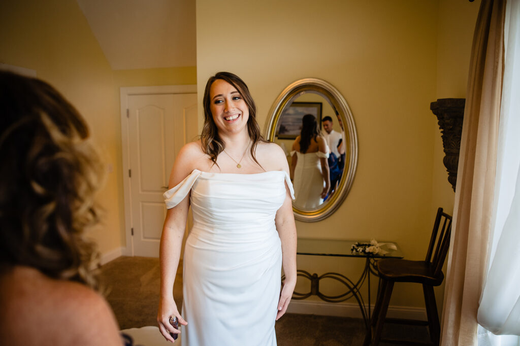 A bride, seen from the back, smiles at her reflection in an ornate mirror, wearing a white off-shoulder wedding dress, with another person reflected in the background.