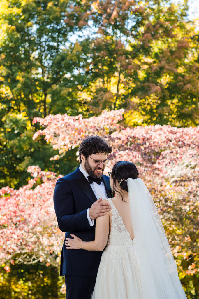 A bride and groom in wedding attire laugh together with autumnal trees in full color in the background.
