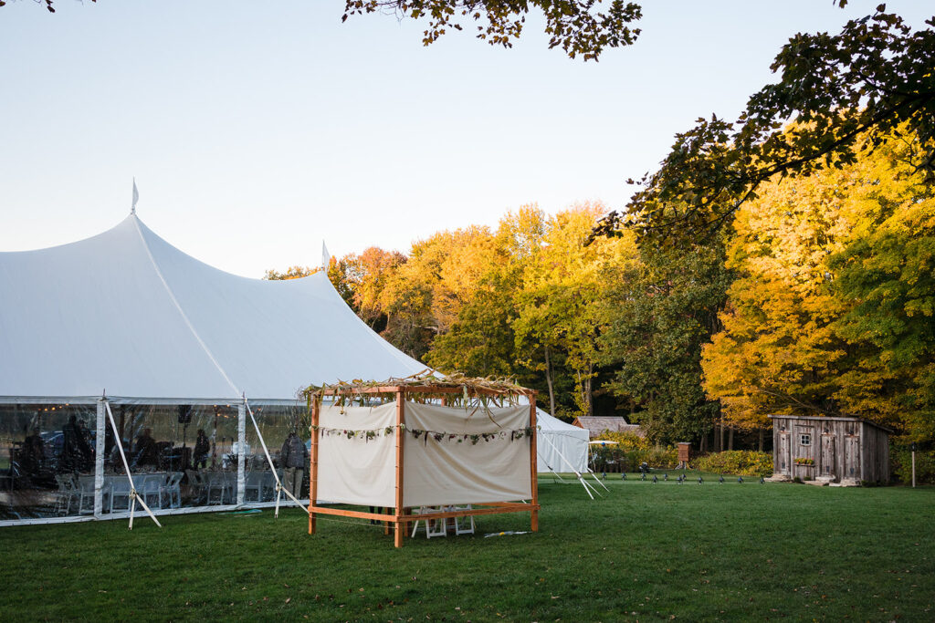 A white wedding tent set up in a field with autumn trees showing yellow foliage in the background during dusk.