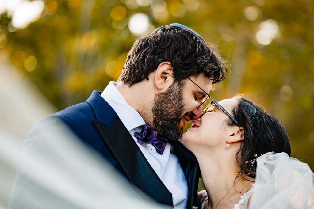 A close-up of a bride and groom touching foreheads lovingly, with a blurred background of autumn leaves.