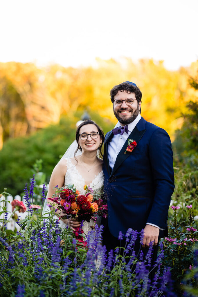A bride and groom standing amidst tall purple flowers, with the bride holding a vibrant bouquet and both smiling at the camera.