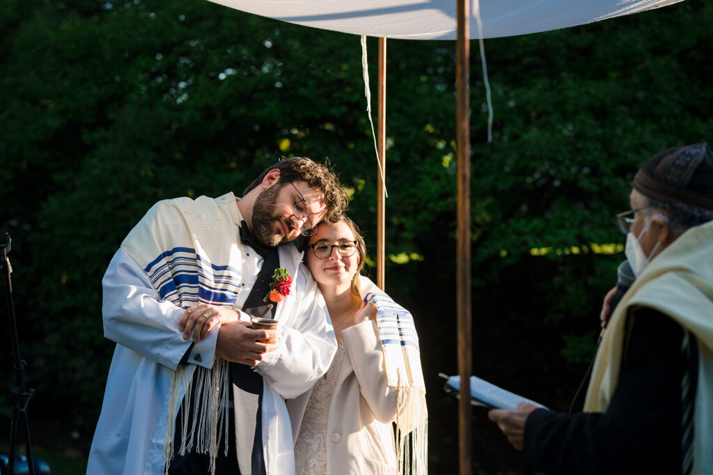 A bride and groom under a chuppah during a Jewish wedding ceremony, with the groom holding a cup and the bride smiling, as an officiant reads from a book.