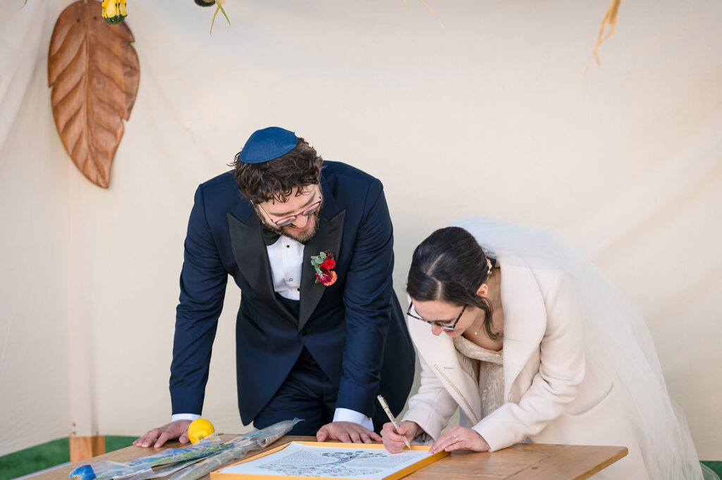 A bride and groom lean over a guest book on a table, smiling as they sign it, with a lemon and blue cloth underneath.