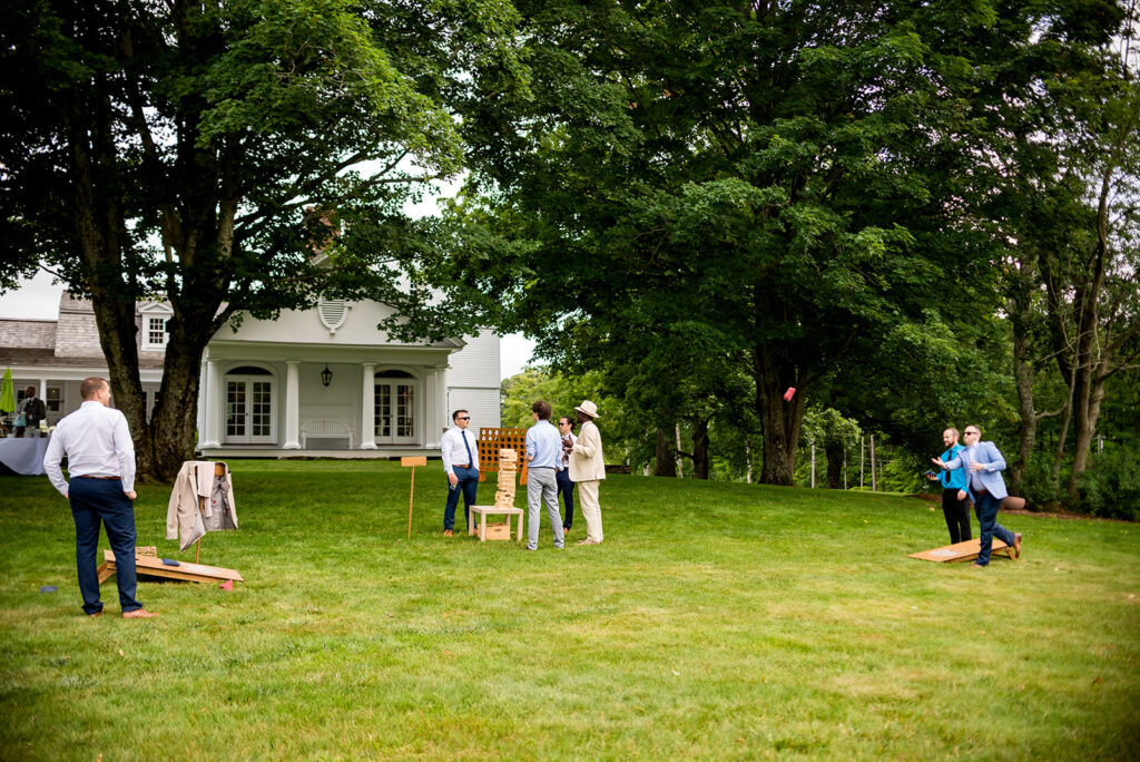 Guests playing a game of cornhole on a lush green lawn with a white colonial house in the background.
