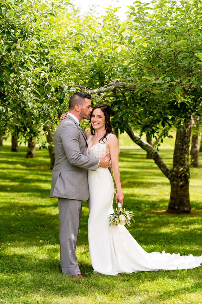 A bride and groom share a tender moment among the green foliage of an apple orchard, with the bride holding a small bouquet.