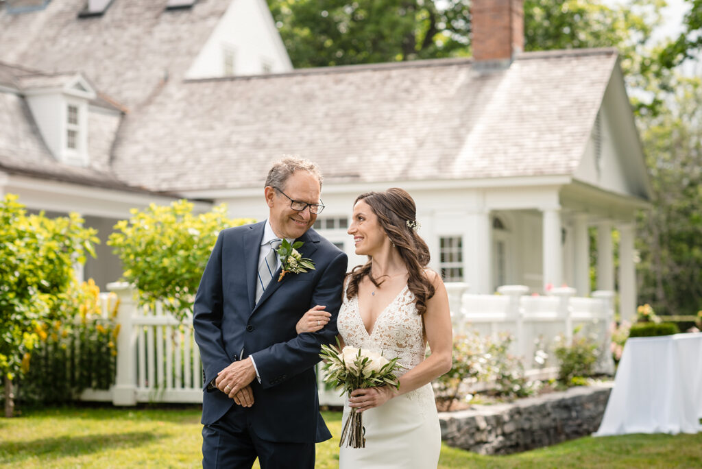 A bride looking up at her father, both smiling, with a well-manicured garden and a white colonial house behind them.