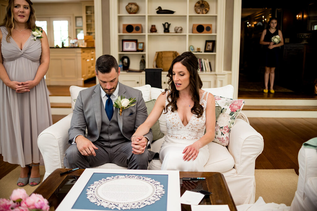 A bride and groom sit on a white sofa signing a Ketubah, with a bridesmaid standing behind them.