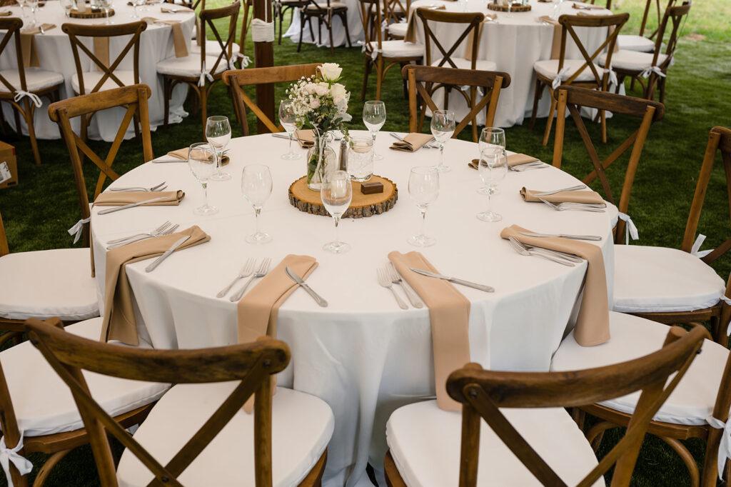 A round wedding table set with white linens, beige napkins, and a wood slice centerpiece with a bouquet of flowers.