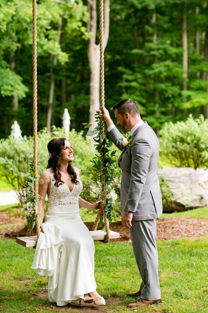 A bride seated on a decorated swing looking up at her groom standing beside her in a garden setting.
