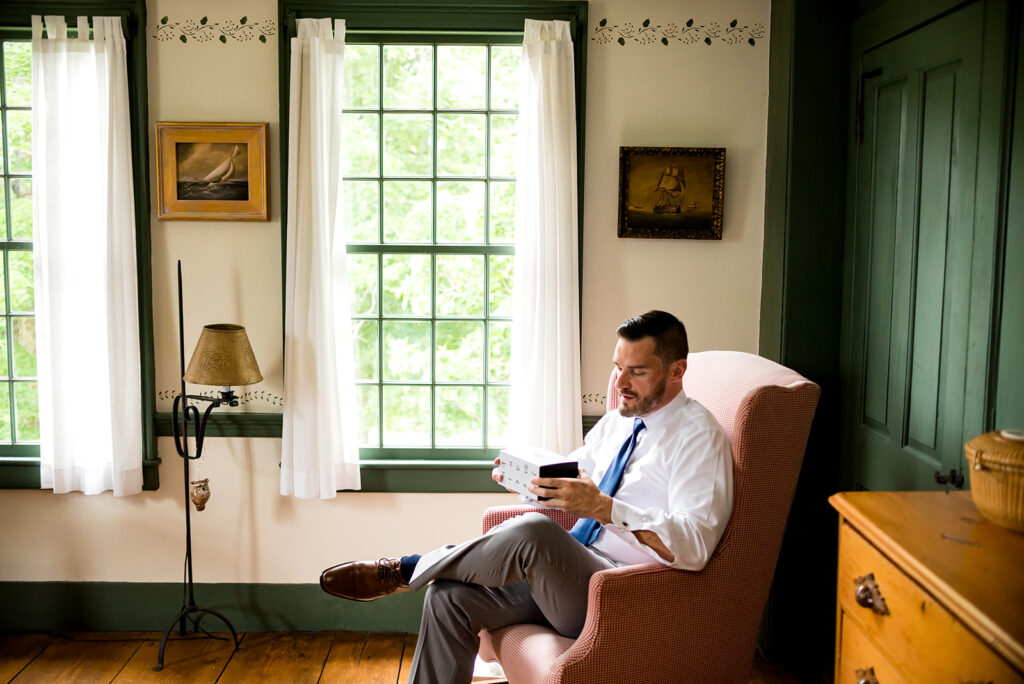 A groom in a blue suit seated in a wingback chair reading a book in a room with traditional decor and green walls.