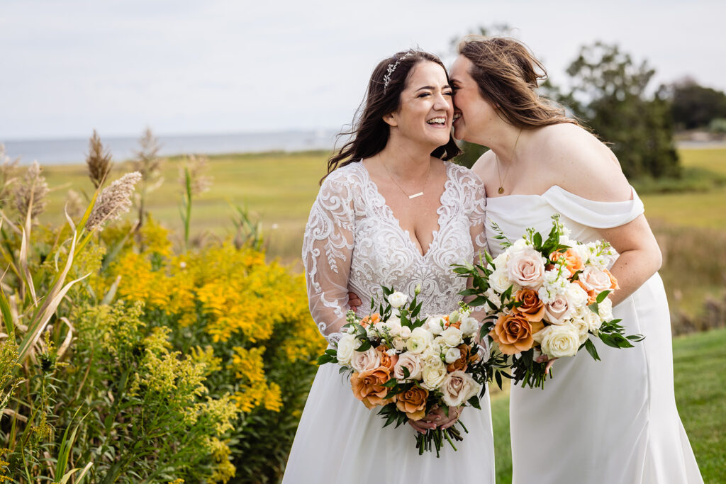 Two brides in white dresses, one with a lace top and the other off-shoulder, share a tender moment among tall grass and wildflowers, with the ocean landscape behind them.