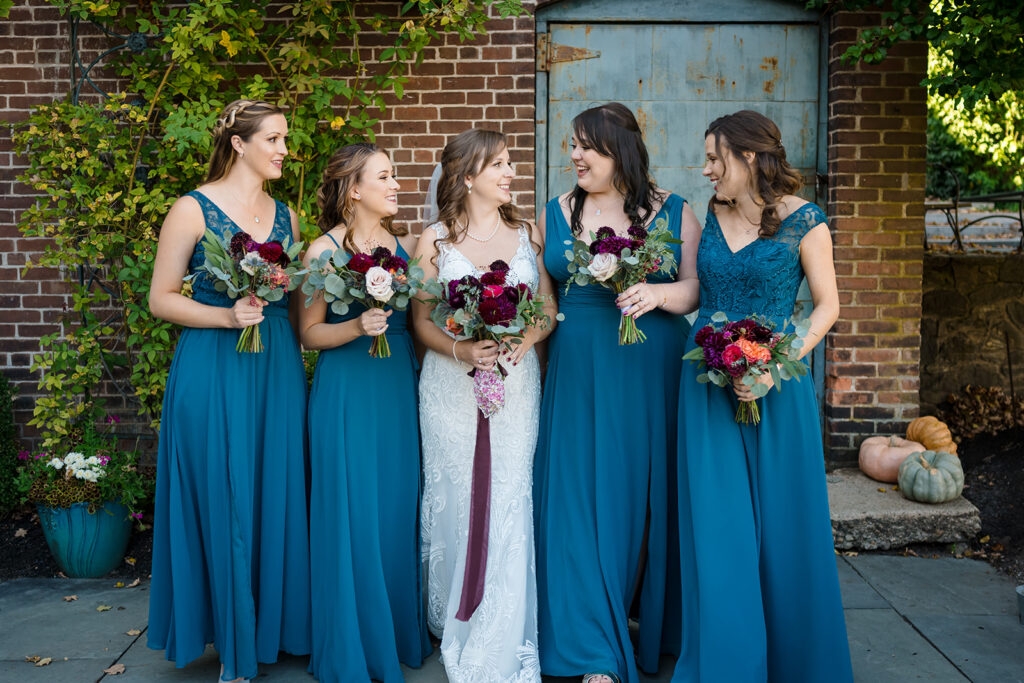 A group of five women in teal bridesmaid dresses with lace details, holding bouquets with deep red and blush flowers, stand beside a bride in a white lace gown.
