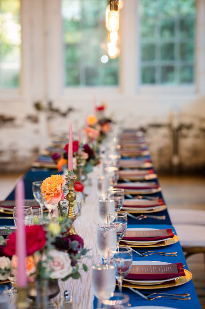 A close-up of a long table with a blue tablecloth, decorated with orange and red flowers, candles, and elegant tableware, with a warm light hanging above.