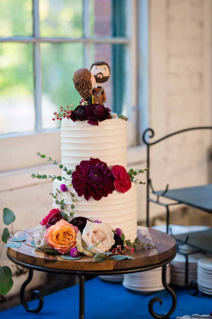 A three-tiered wedding cake adorned with deep red and blush flowers, topped with a bride and groom figurine, set against a bright window.