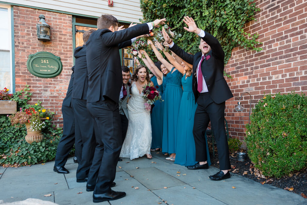 A jubilant wedding party raising their arms in celebration around the bride and groom outside a brick building with ivy.