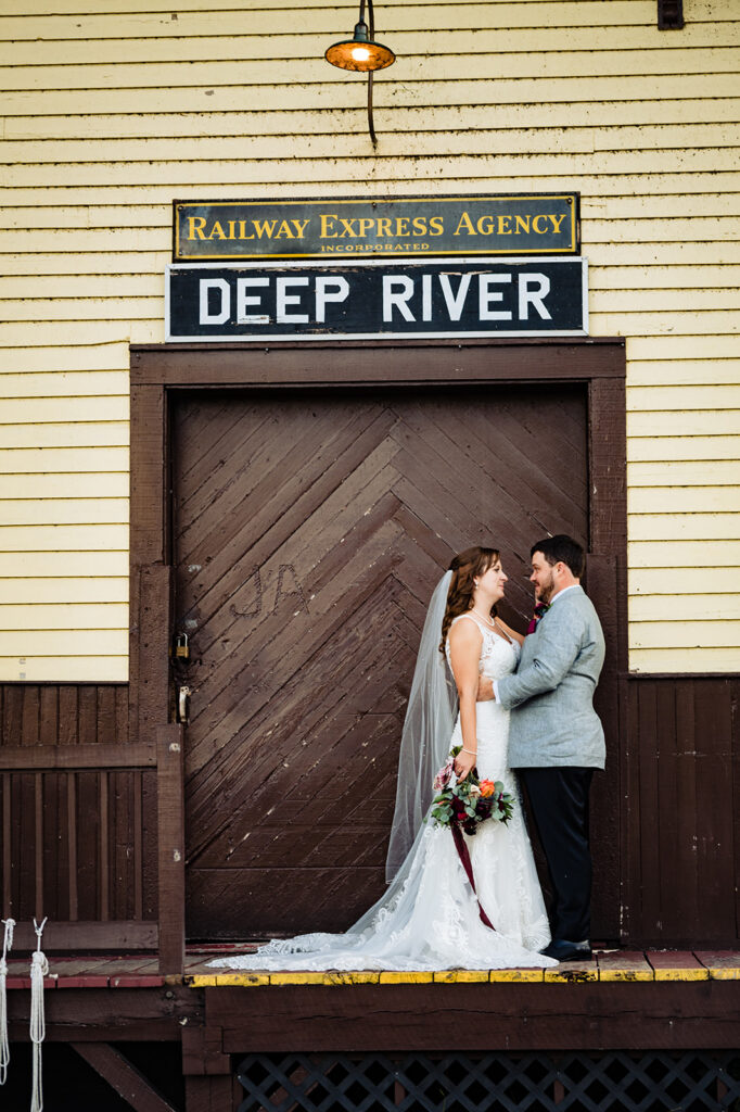 A bride and groom share a moment in front of a rustic brown door labeled "Railway Express Agency DEEP RIVER," with a vintage light fixture above them.