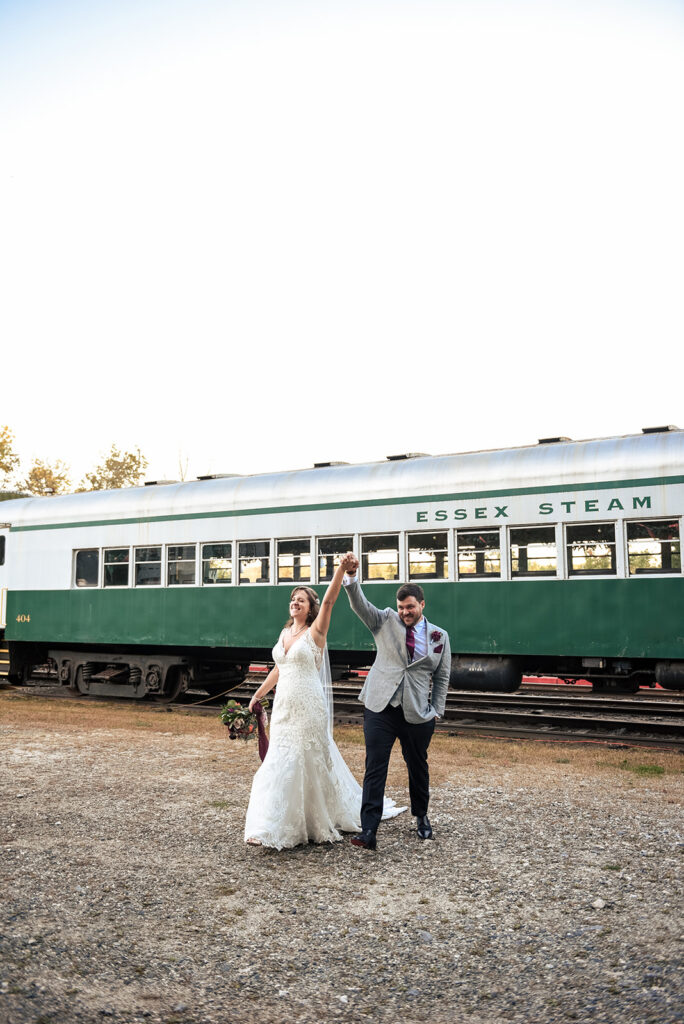 The bride and groom joyfully dance in front of a green vintage train car labeled "ESSEX STEAM," under a clear blue sky.