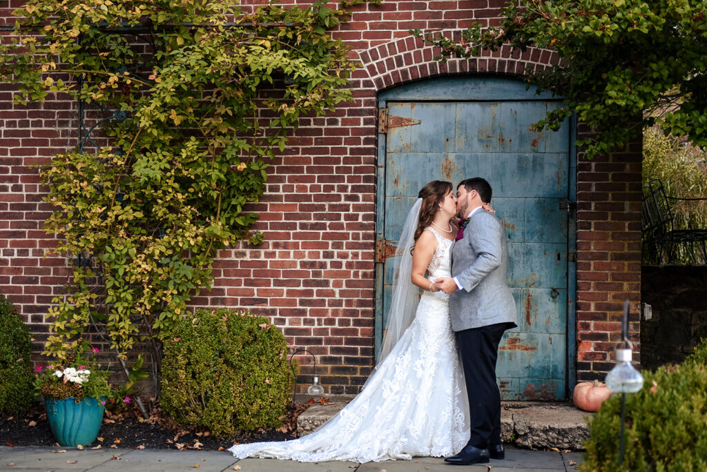 A couple in wedding attire share a kiss by a weathered blue door, surrounded by brick walls and autumnal plants.