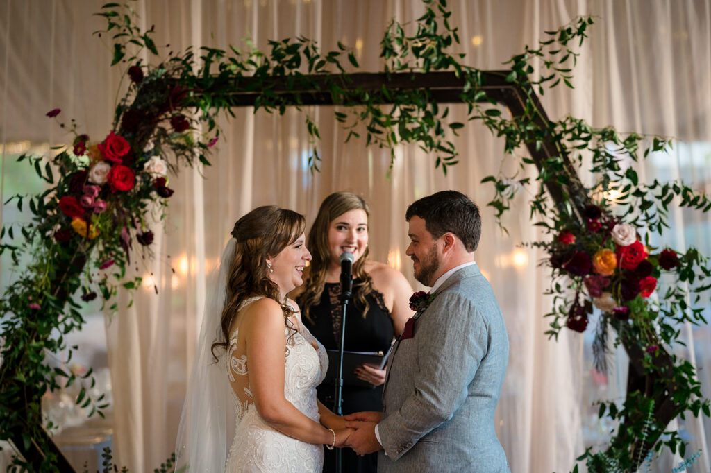 An indoor wedding ceremony with a couple holding hands at the altar, a black-dressed officiant behind them, and a floral arch.