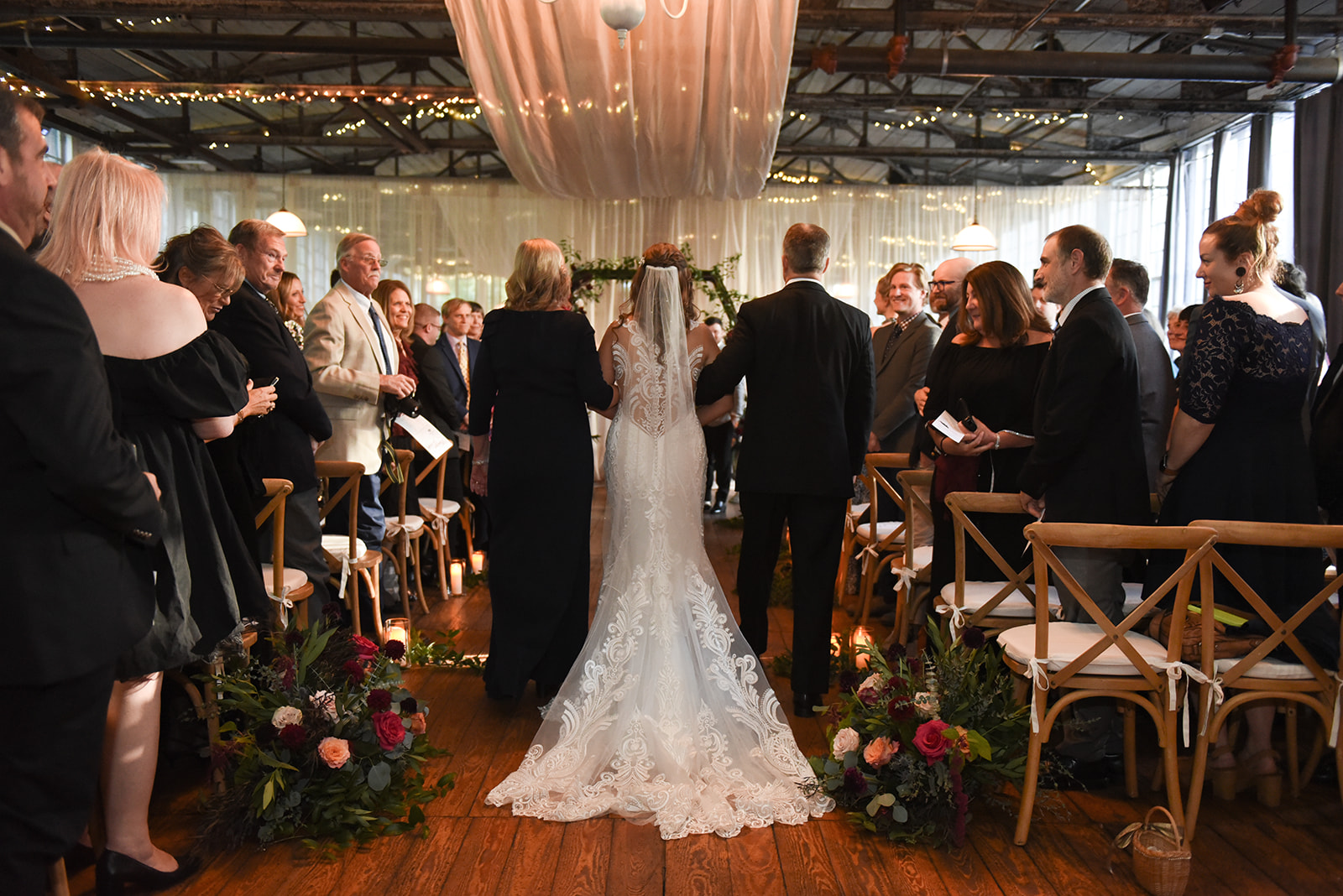 The back view of a bride walking down the aisle in a lace dress, guests on either side seated on folding chairs, and a floral-lined aisle leading to a draped fabric backdrop.