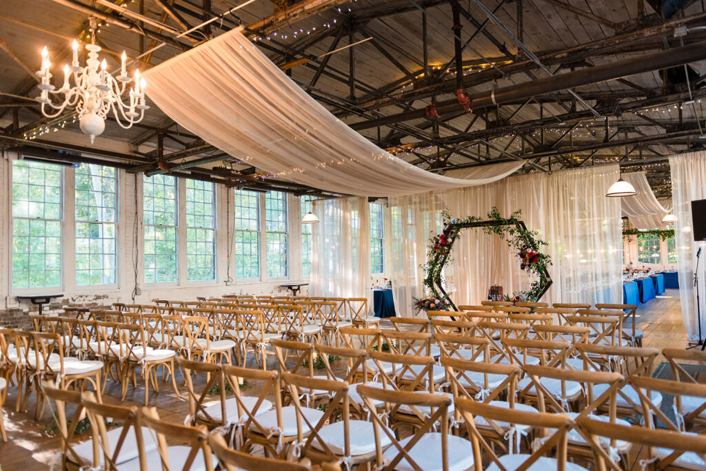 A spacious wedding venue interior with wooden floors, draped ceiling fabric, a floral arch, and rows of wooden chairs before a large window.