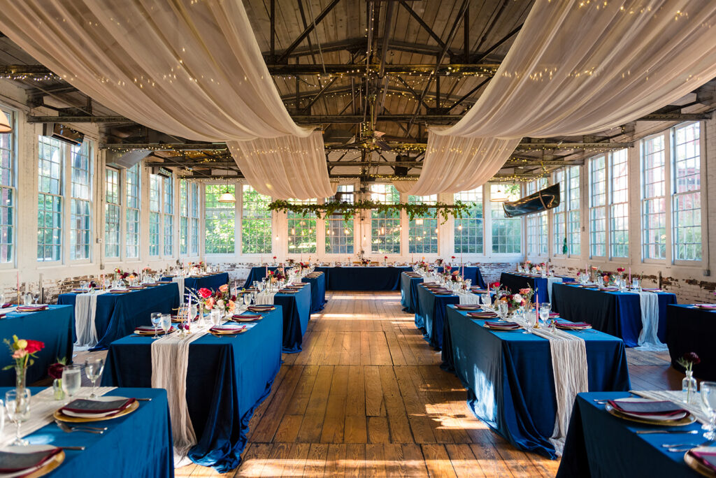 A grand wedding reception hall with wooden floors, blue tablecloths, and rustic chandeliers, set for a celebratory meal.