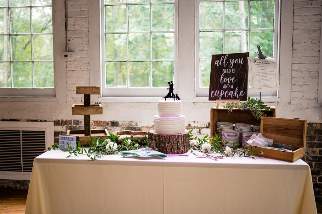 A rustic wedding dessert table with a cake on a wooden stump stand, a sign reading "All you need is love and a cupcake," and a display of coffee essentials by a large window.