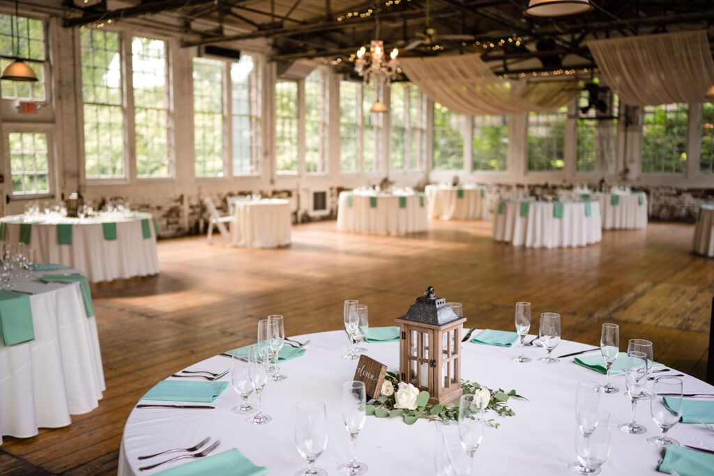 An elegant wedding reception hall with round tables set with white linens, teal napkins, and simple centerpieces, in a spacious room with large windows and draping white fabric