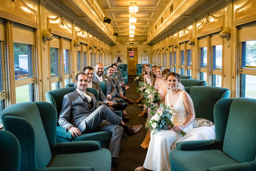 A wedding party seated inside a vintage train car, with the groomsmen and bridesmaids surrounding the smiling bride holding a bouquet