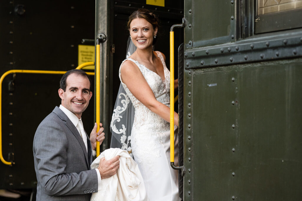 A bride in a lace gown and a groom in a gray suit smiling at the entrance of a vintage train car.