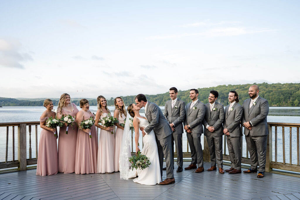 A bride and groom share a kiss on a lakeside deck with bridesmaids in blush dresses and groomsmen in gray suits to the side, overlooking a serene body of water.