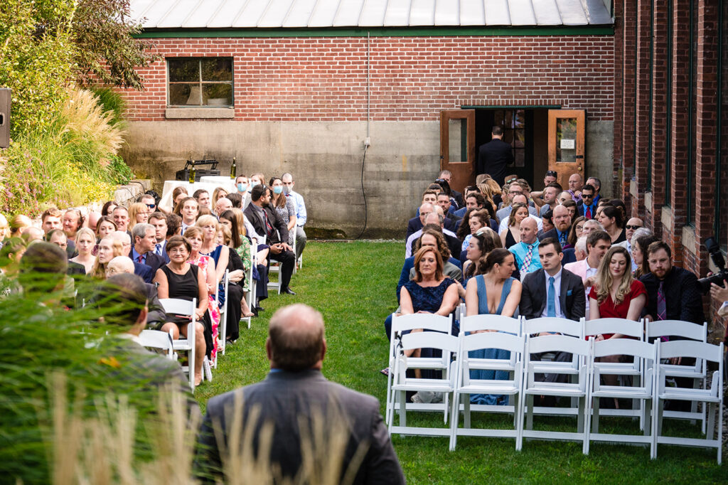 Wedding guests seated on white chairs during an outdoor ceremony, facing an aisle with a view of the officiant in the background.