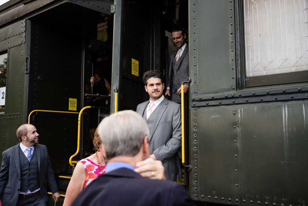 Groomsmen standing at the open door of a vintage train, looking towards the guests outside.