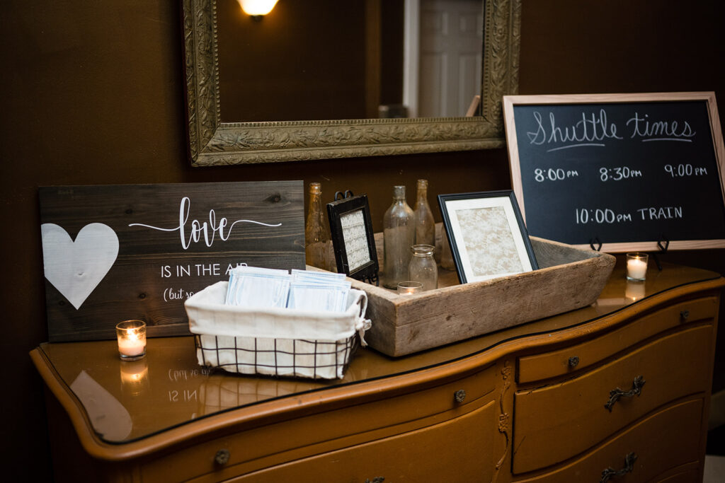 A vintage wooden table holds wedding reception items, including a sign saying "Love is in the air," a schedule of shuttle times, and a basket of face masks.