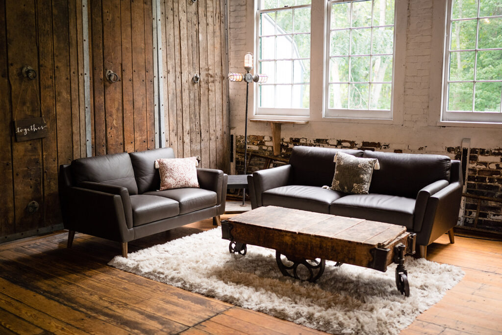 A cozy rustic lounge area with two dark leather sofas, a wooden coffee table on wheels, set against an old wooden wall with large windows
