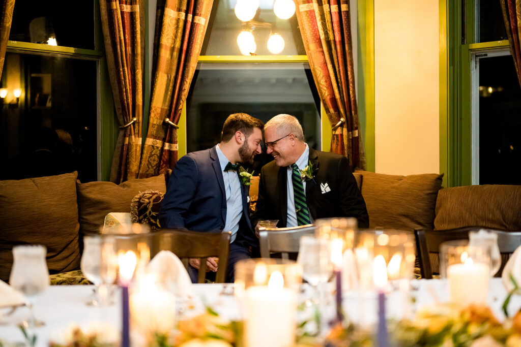 Two grooms leaning in for a sweet moment, one in a blue suit and the other in black, both with green boutonnieres, in a warmly lit vintage room
