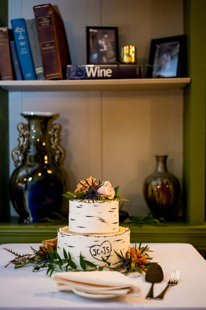 A two-tiered wedding cake with white birch bark texture and floral decorations on top, displayed on a table with books and a vase in the background