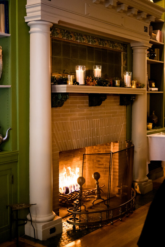 A romantic and warmly lit fireplace with candles on the mantel, surrounded by decorative tiles and a classical mirror reflecting a wedding setting