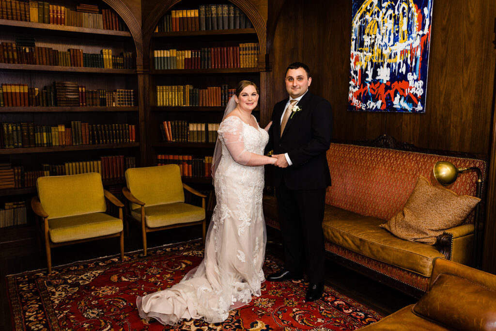 A bride and groom holding hands in a grand library, surrounded by shelves of classic books, with a vibrant abstract painting in the background, sharing a moment of unity.