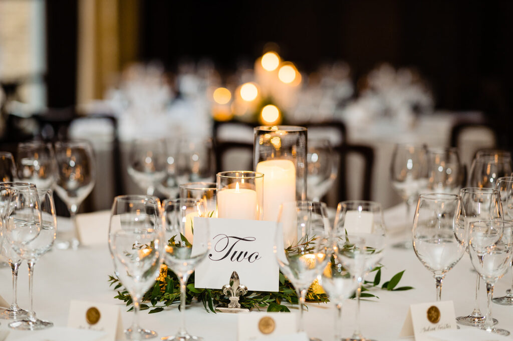 Elegant wedding table setting with 'Two' table number card, surrounded by wine glasses, candles, and greenery