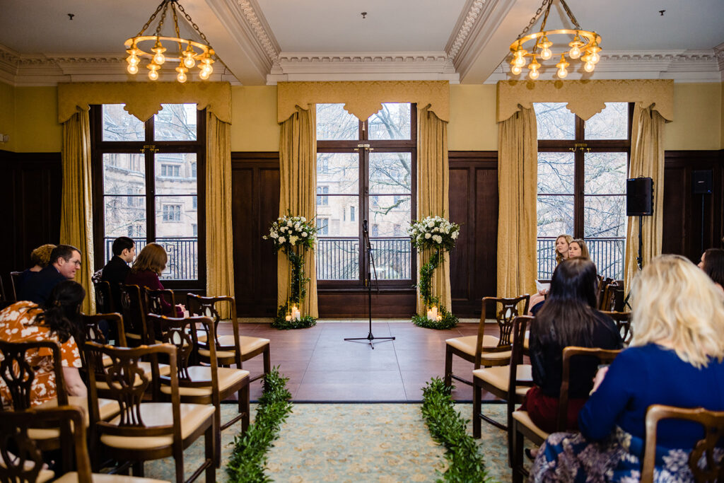 Intimate wedding ceremony setup with floral arrangements and candles in a historic room with large windows and ornate chandeliers.
