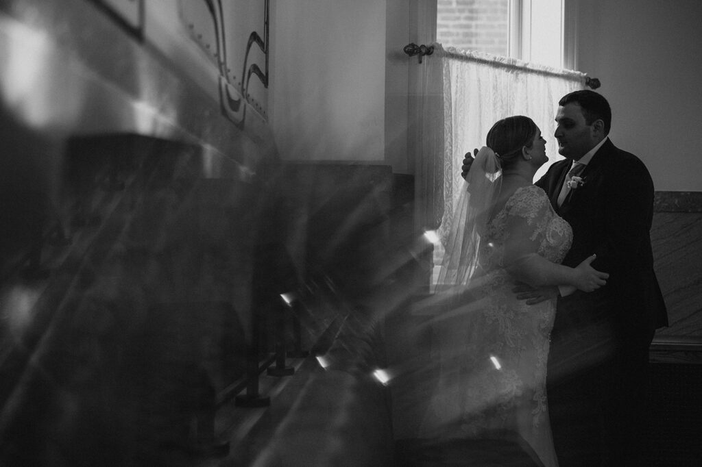 A romantic black and white photograph capturing a bride and groom embracing, with light flares and a sheer curtain adding a dreamy effect