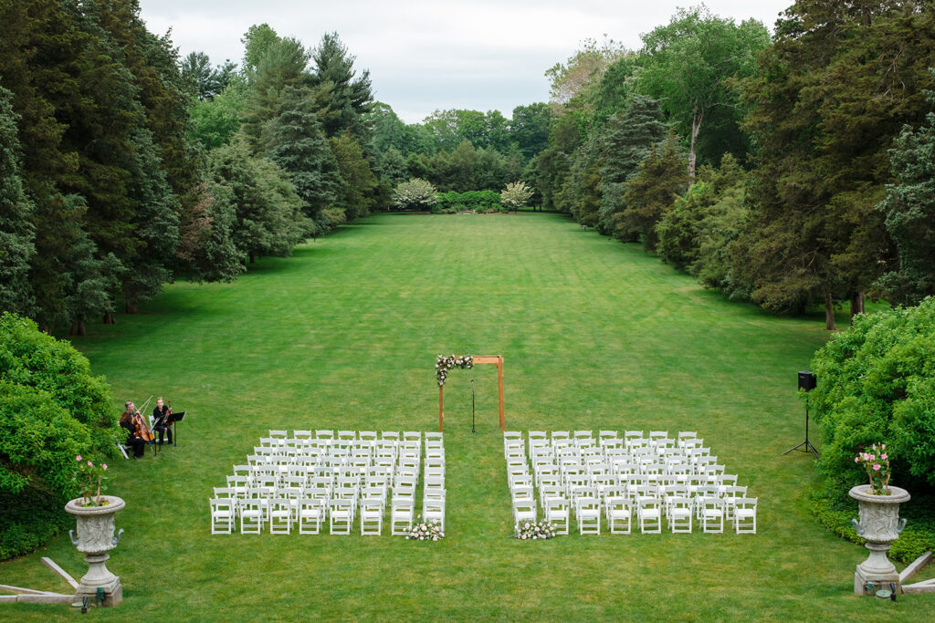 Aerial view of an outdoor wedding ceremony setup at Wadsworth Mansion, with rows of white chairs facing a floral arch, flanked by trees