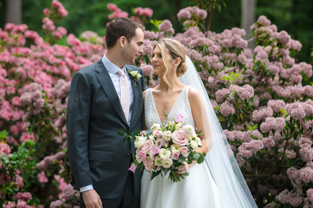 Close-up of a smiling bride and groom with a pink and white floral bouquet, standing in front of blooming pink hydrangeas.